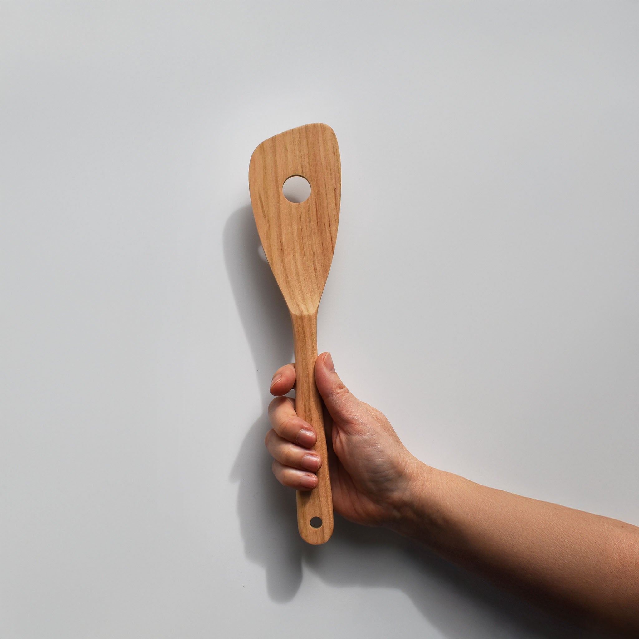 Sakura wood slotted spatula with hanging hole and natural wood grain held by hand for scale