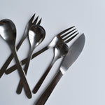 A set of Sori Yanagi stainless steel cutlery, including knives, forks, and spoons, arranged on a white surface, showcasing minimalist Japanese design and high-quality craftsmanship.