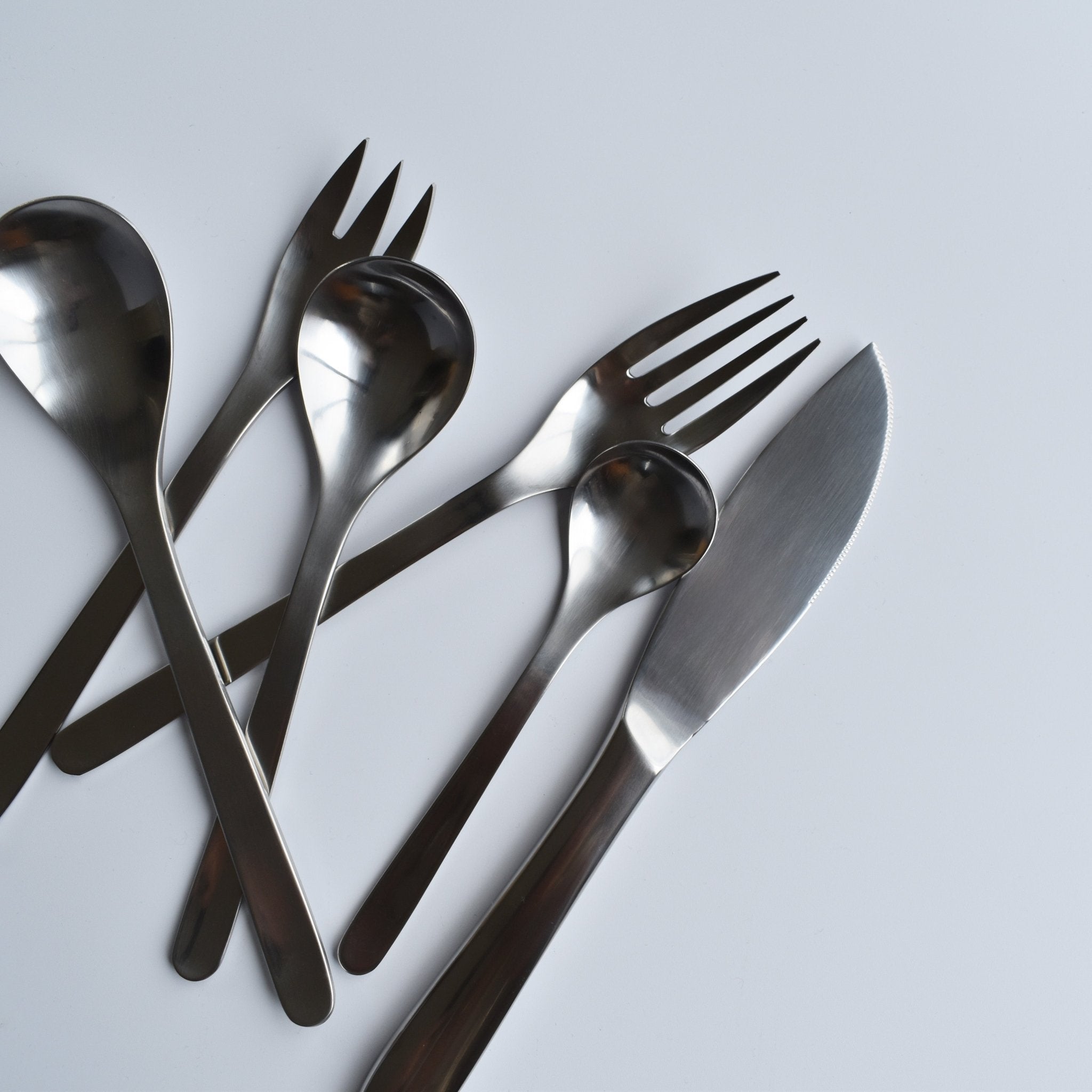 A set of Sori Yanagi stainless steel cutlery, including knives, forks, and spoons, arranged on a white surface, showcasing minimalist Japanese design and high-quality craftsmanship.