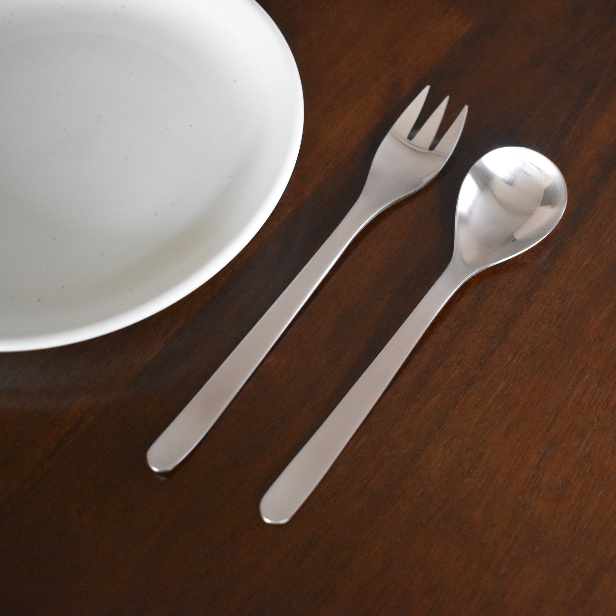 Sori Yanagi stainless steel fork and spoon placed beside a white ceramic plate on a wooden dining table, highlighting minimalist Japanese tableware design.