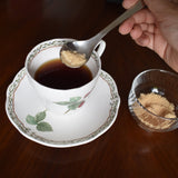 A Sori Yanagi stainless steel tea spoon adding brown sugar to a cup of black tea on a wooden table, with a small glass bowl of sugar nearby.