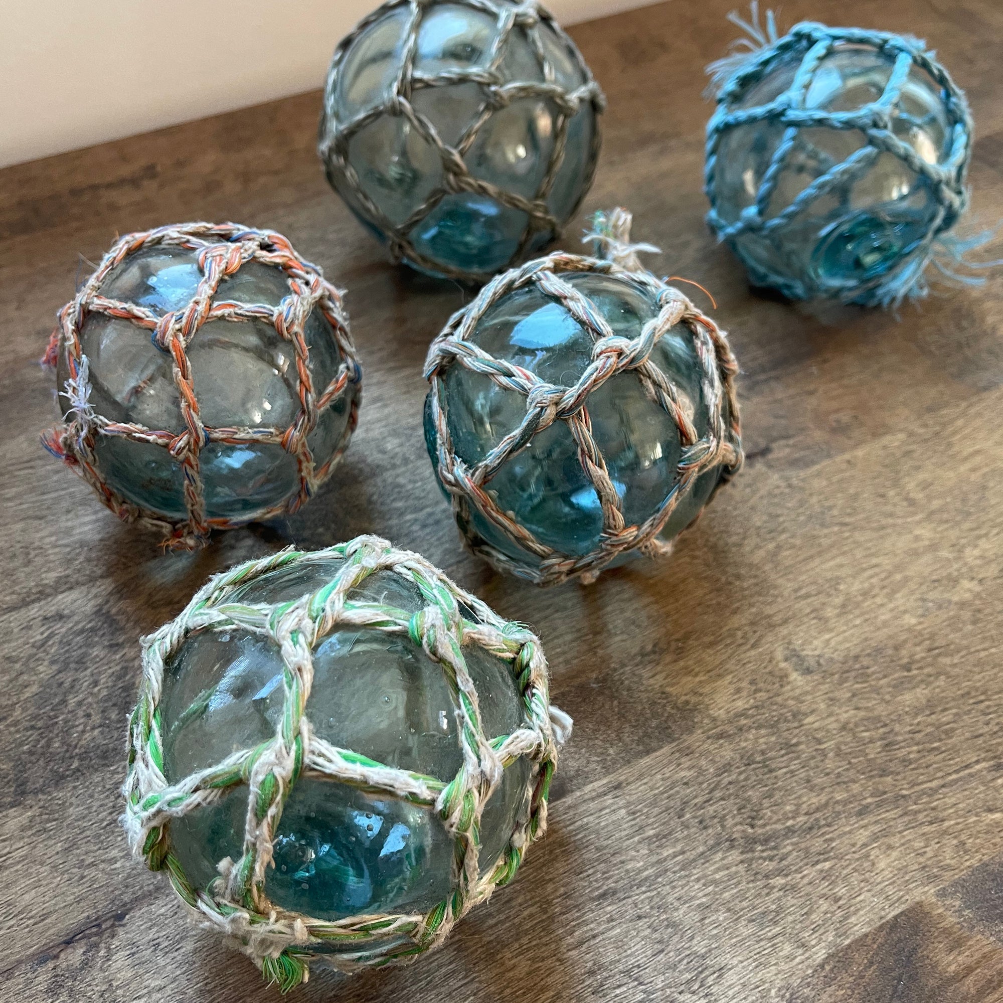 A collection of vintage Japanese glass fishing floats in hand-knotted multicolor rope nets, showcasing rustic coastal decor pieces with individual character and patina, displayed on a warm wood surface.