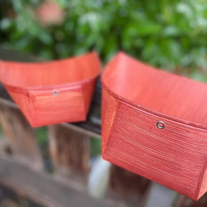 Two red Harimi dustpans made of persimmon-coated washi paper resting on a wooden railing, showcasing their curved design and traditional Japanese craftsmanship