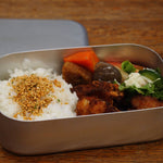 Close-up of a Japanese bento box filled with rice, karaage chicken, and assorted vegetables inside a sleek aluminum container on a wooden surface.