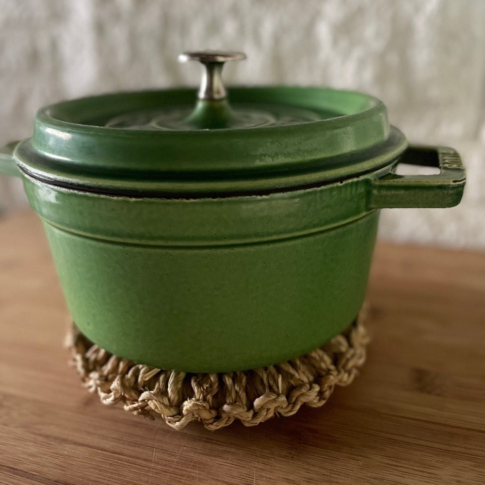 Green cast iron pot resting on a round handwoven Japanese straw trivet atop a wooden surface, showing protection from heat and natural kitchen style