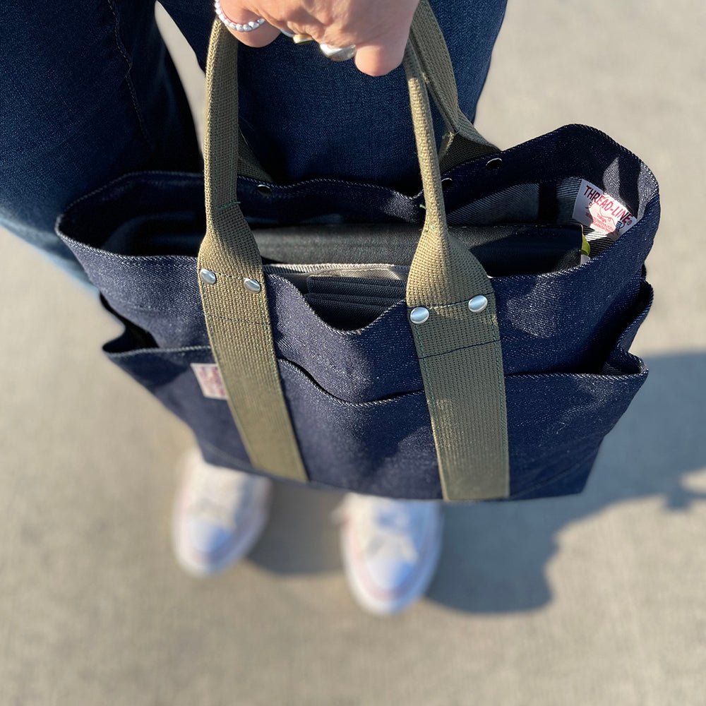 Top-down view of a person holding a compact Japanese denim tote bag with olive handles, showing outer pockets and organized contents inside