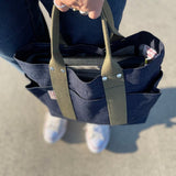 Top-down view of a person holding a compact Japanese denim tote bag with olive handles, showing outer pockets and organized contents inside