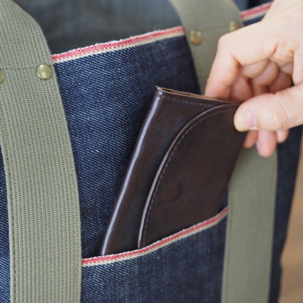 Hand placing brown wallet into outer pocket of Japanese selvage denim tote bag with red stitching and olive green handles