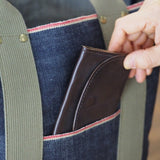 Hand placing brown wallet into outer pocket of Japanese selvage denim tote bag with red stitching and olive green handles