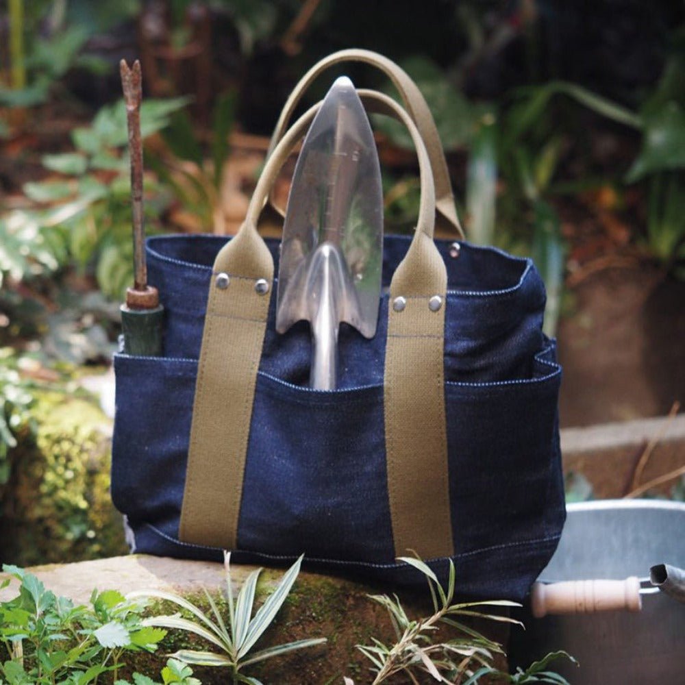 Denim tote bag with olive handles used as a garden tool bag, holding a trowel and weeder, shown outdoors in a lush garden setting