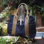 Denim tote bag with olive handles used as a garden tool bag, holding a trowel and weeder, shown outdoors in a lush garden setting