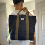 Back view of a person holding a Japanese selvage denim tote bag with olive webbing handles and visible Thread Line Tokyo label indoors