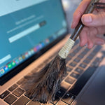 Hand using a mini feather duster to clean a laptop keyboard, highlighting its precision for delicate electronics.