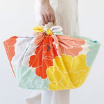 A hand carrying a basket wrapped in a colorful furoshiki cloth featuring a vibrant peony floral design in orange, yellow, mint green, peach, and lavender. The fabric is neatly tied at the top with visible floral petals creating a cheerful, eco-friendly alternative to a traditional bag.