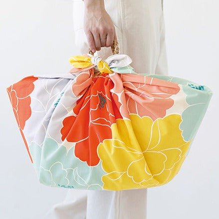 A hand carrying a basket wrapped in a colorful furoshiki cloth featuring a vibrant peony floral design in orange, yellow, mint green, peach, and lavender. The fabric is neatly tied at the top with visible floral petals creating a cheerful, eco-friendly alternative to a traditional bag.