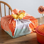 A square box wrapped in a vibrant peony-patterned furoshiki cloth, showcasing hues of orange, mint green, lavender, and peach with a contrasting yellow bow on top. The wrapped box sits on a wooden dining table beside a red-orange cast iron pot and a single flower in a glass vase, evoking a cozy and thoughtful presentation style.