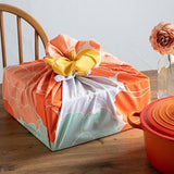 A square box wrapped in a vibrant peony-patterned furoshiki cloth, showcasing hues of orange, mint green, lavender, and peach with a contrasting yellow bow on top. The wrapped box sits on a wooden dining table beside a red-orange cast iron pot and a single flower in a glass vase, evoking a cozy and thoughtful presentation style.