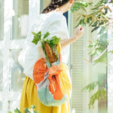 A woman walking outdoors in natural sunlight carries groceries wrapped in a colorful orange, mint green, and yellow peony-patterned furoshiki bag tied in the traditional Hime Musubi style, with a baguette and leafy greens peeking out from the top.