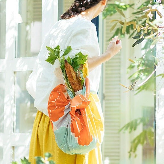 A woman walking outdoors in natural sunlight carries groceries wrapped in a colorful orange, mint green, and yellow peony-patterned furoshiki bag tied in the traditional Hime Musubi style, with a baguette and leafy greens peeking out from the top.