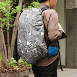 A person walking outdoors with a large backpack completely wrapped in a khaki ISA-MONYO “knot” patterned furoshiki. The dynamic black-and-cream swirl design covers the entire bag, showcasing the furoshiki’s adaptability as a modern, water-repellent backpack cover for everyday urban use.