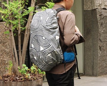 A person walking outdoors with a large backpack completely wrapped in a khaki ISA-MONYO “knot” patterned furoshiki. The dynamic black-and-cream swirl design covers the entire bag, showcasing the furoshiki’s adaptability as a modern, water-repellent backpack cover for everyday urban use.