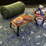 A picnic setup featuring a green sleeping bag, a mini food tray with sandwiches, cups of soup, and a wicker basket filled with snacks—all placed atop a furoshiki cloth with the khaki ISA-MONYO “knot” design. The swirling black and cream line pattern adds a modern touch to the outdoor scene, making the furoshiki serve as both a groundsheet and a stylish element of the picnic.