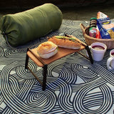 A picnic setup featuring a green sleeping bag, a mini food tray with sandwiches, cups of soup, and a wicker basket filled with snacks—all placed atop a furoshiki cloth with the khaki ISA-MONYO “knot” design. The swirling black and cream line pattern adds a modern touch to the outdoor scene, making the furoshiki serve as both a groundsheet and a stylish element of the picnic.