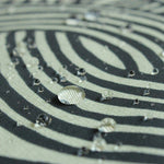 A close-up shot showing water droplets beading on the surface of a water-repellent furoshiki cloth. The fabric features the ISA-MONYO “knot” design in khaki, with black and cream-colored interwoven line patterns. The image highlights the fabric’s water-resistant properties, as the droplets sit cleanly on top of the tightly woven material without soaking in.