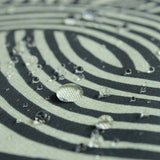 A close-up shot showing water droplets beading on the surface of a water-repellent furoshiki cloth. The fabric features the ISA-MONYO “knot” design in khaki, with black and cream-colored interwoven line patterns. The image highlights the fabric’s water-resistant properties, as the droplets sit cleanly on top of the tightly woven material without soaking in.