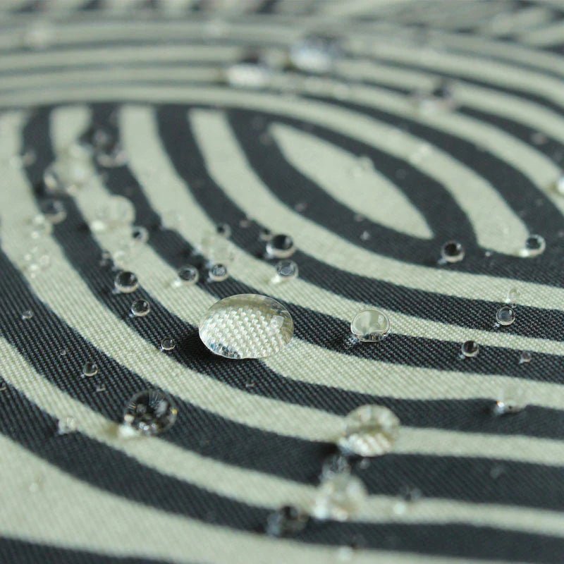 A close-up shot showing water droplets beading on the surface of a water-repellent furoshiki cloth. The fabric features the ISA-MONYO “knot” design in khaki, with black and cream-colored interwoven line patterns. The image highlights the fabric’s water-resistant properties, as the droplets sit cleanly on top of the tightly woven material without soaking in.