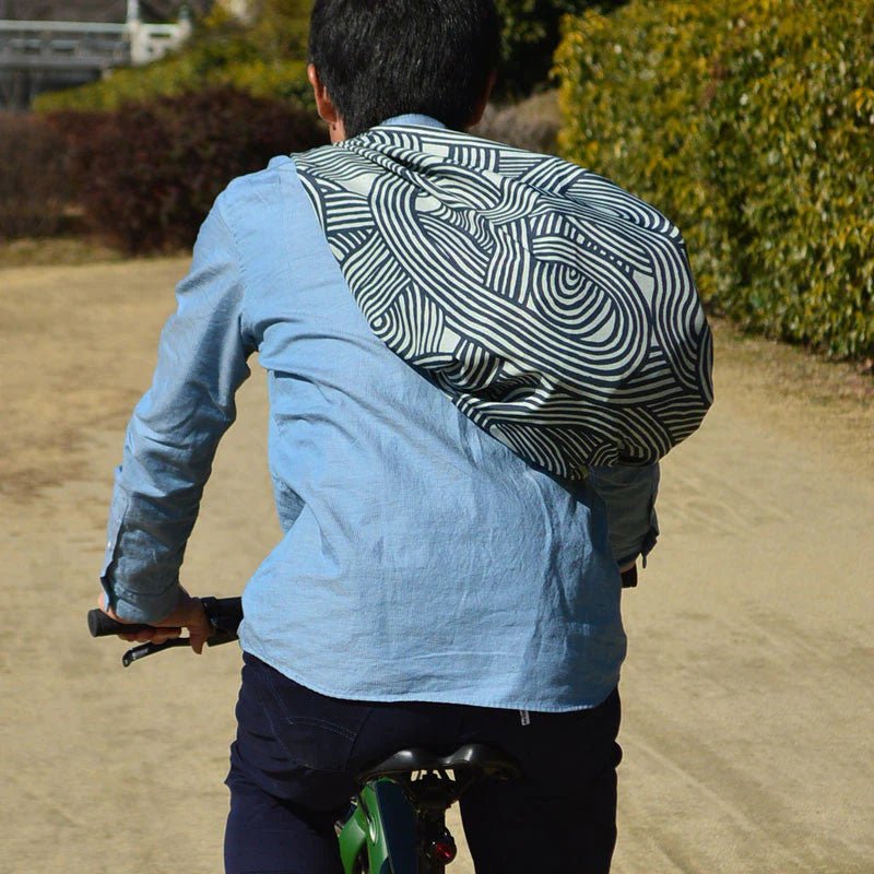 A person riding a bicycle with a large furoshiki cloth tied diagonally across their back like a sling bag. The cloth features a bold, modern khaki and white abstract pattern from the ISA-MONYO series. The scene is outdoors on a dirt path surrounded by greenery, and the rider is wearing a light blue button-up shirt and dark pants. The furoshiki is styled to function as a practical and stylish carrying solution for commuting or casual biking.