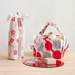 Japanese furoshiki gift wraps on a wooden table—one bottle wrapped in floral fabric, one box wrapped in Yumeji Takehisa’s Tsunagi Dango pattern, paired with wine glasses and gift box for a festive display