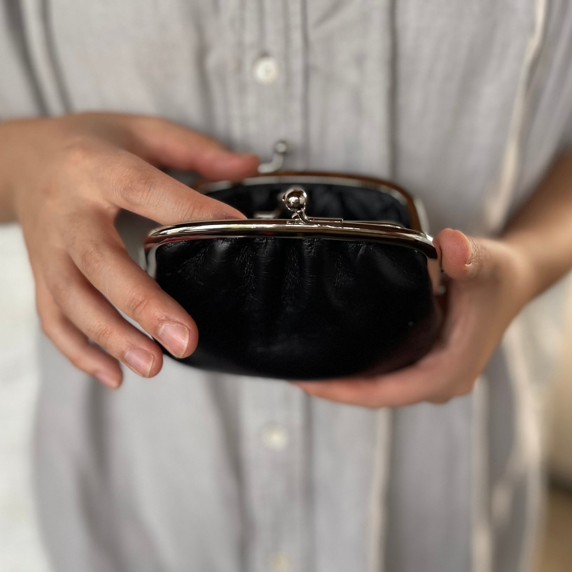 Black leather Gamaguchi purse held in both hands, showing the compact size, silver clasp, and ease of use in everyday life.