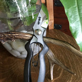 Hidehisa garden pruners resting on a deer antler with a glass vase of white stones and greenery in the background, showcasing Japanese craftsmanship in a rustic indoor setting