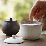 Hand reaching into a handcrafted white Shigaraki ceramic salt jar filled with salt, with a matching lid placed beside it and a dark charcoal version in the background, set on a wooden table.