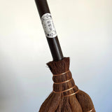 Detailed shot of the handle of a traditional Japanese shuro angle broom, featuring a “Kaneichi” brand label and tightly bound copper wire where the palm fibers meet the handle.