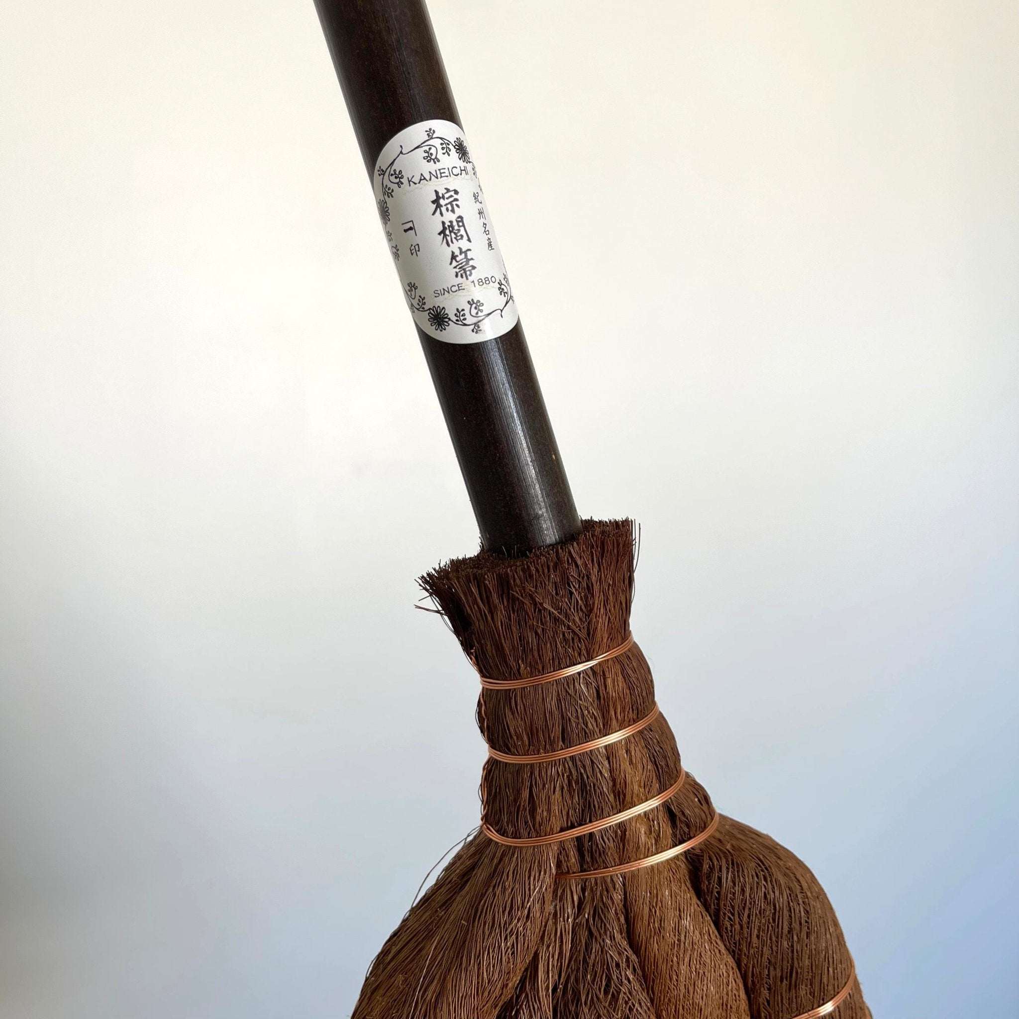 Detailed shot of the handle of a traditional Japanese shuro angle broom, featuring a “Kaneichi” brand label and tightly bound copper wire where the palm fibers meet the handle.
