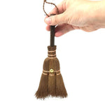 A hand holding a small Japanese shuro whisk broom by its dark wooden handle, showing its compact size, natural palm fiber bristles, and copper wire binding.