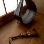 A small Japanese shuro whisk broom with copper bindings resting on a wooden floor beside a sculptural dark dustpan, placed near a shoji screen for a serene, traditional ambiance.