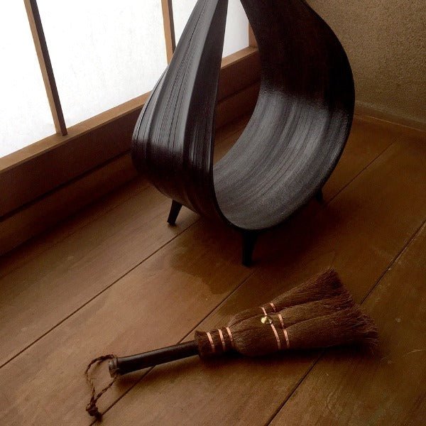 A small Japanese shuro whisk broom with copper bindings resting on a wooden floor beside a sculptural dark dustpan, placed near a shoji screen for a serene, traditional ambiance.