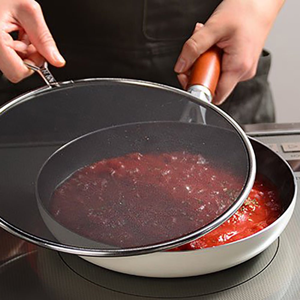 Close-up of a splatter screen being lifted off a frying pan with simmering tomato sauce, showing its use in preventing oil and sauce splatter while cooking